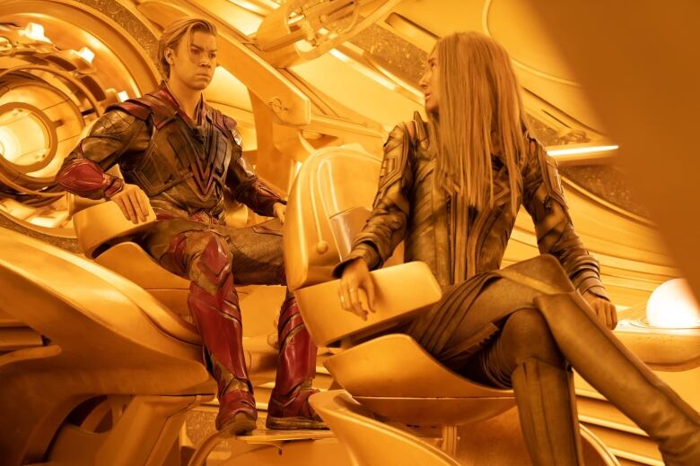 A man and a woman are seated in a futuristic spaceship. The interior of the spaceship is entirely gold-coloured. Both characters have golden skin and gold clothing. The woman seated in the front is looking back at the man seated behind her. Both have concerned looks on their faces. 