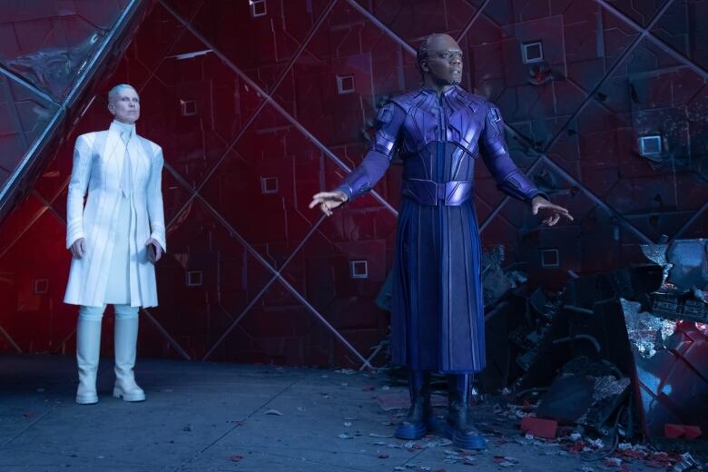 A man in a purple robe, and what appears to be skin artificially stitched onto his head,  stands next to a pile of rubble in a futuristic room. To his right a figure wearing all white robes looks on. 