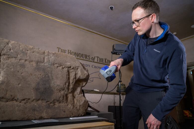 Man in blue sweater holds a piece of equipment scanning an old stone.