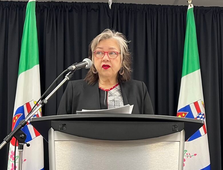 A woman speaks from behind a podium, in front of a backdrop of flags.