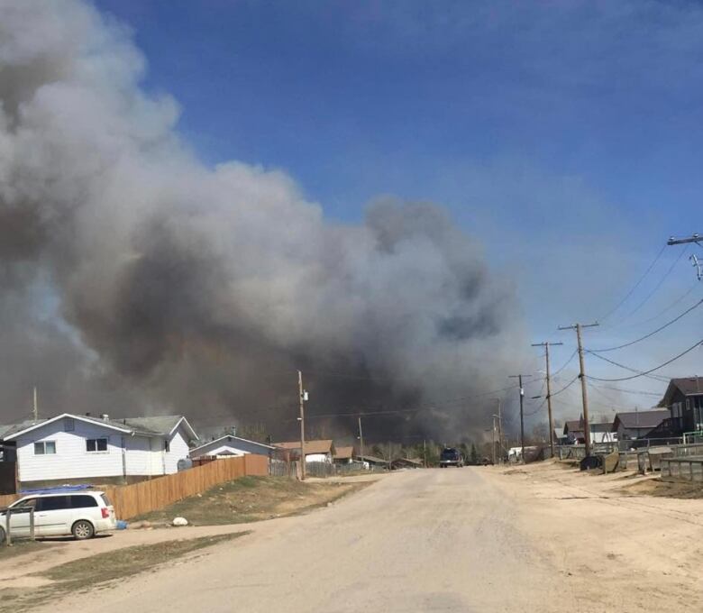 Thick black smoke billows in the distance with a backdrop of clear blue sky. a home and paved road appear in the foreground. 