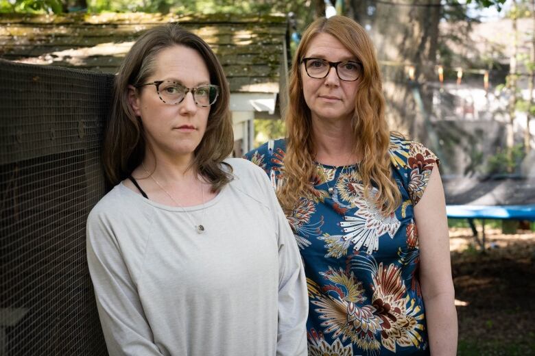 A woman with brown hair and a woman with orange hair look into the camera. Behind them is a wooden shed, a large tree and a trampoline. 