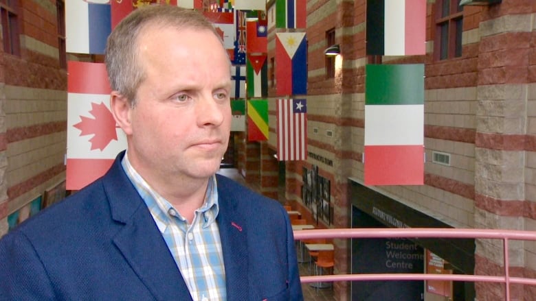 A white man in a blue suit jacket and checked shirt stands in front of an indoor balcony overlooking hanging strands of flags