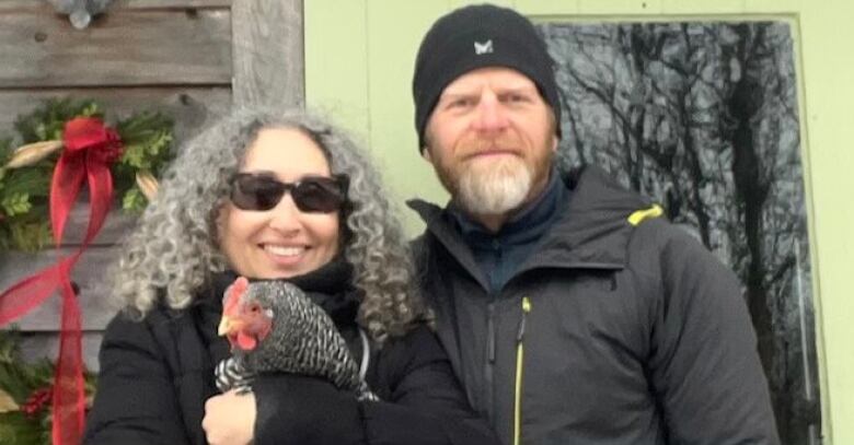 Older man and woman wearing jackets in front of a house with a green wreath and door behind them. Woman is holding a chicken. 