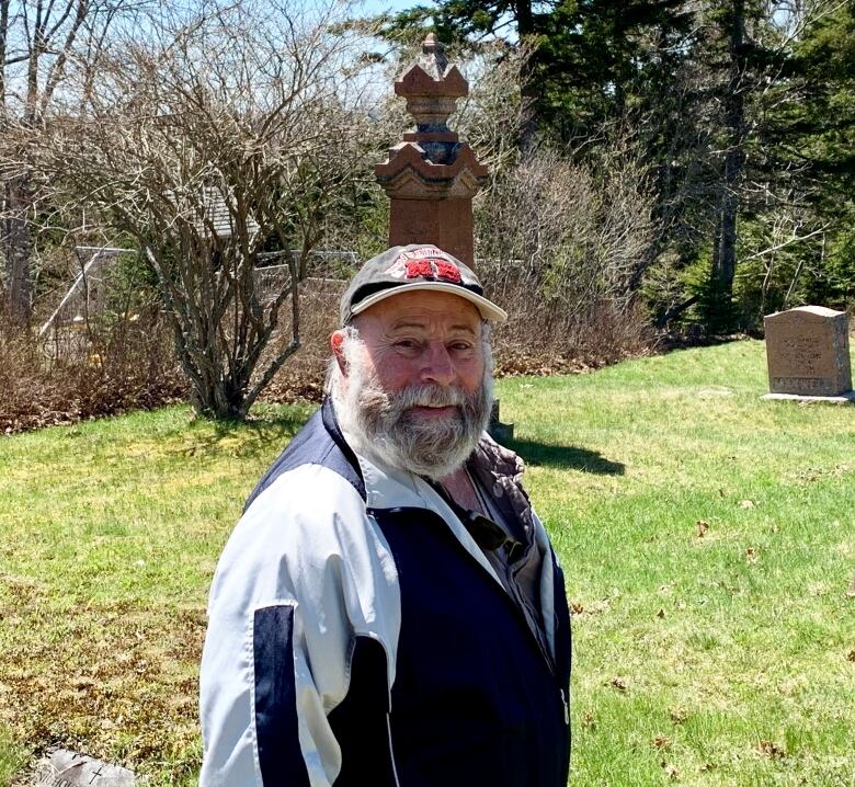 Older man wearing wind breaker jacket and baseball cap standing with his body away and face towards the camera, with grass and gravestones behind him.