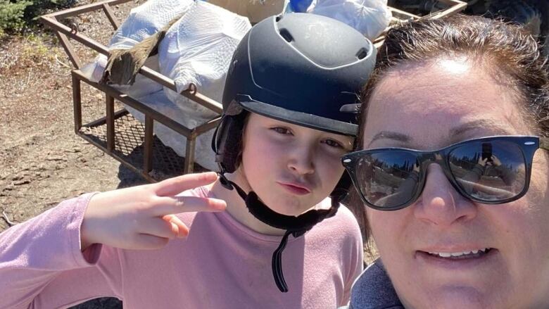 A girl in a helmet makes a duck face at the camera and a peace sign while a woman looks at the camera. Behind them is an ATV and a trailer filled with trash bags. 