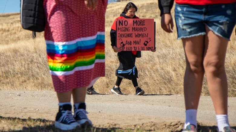 A person in a ribbon skirt holds a sign that says, 