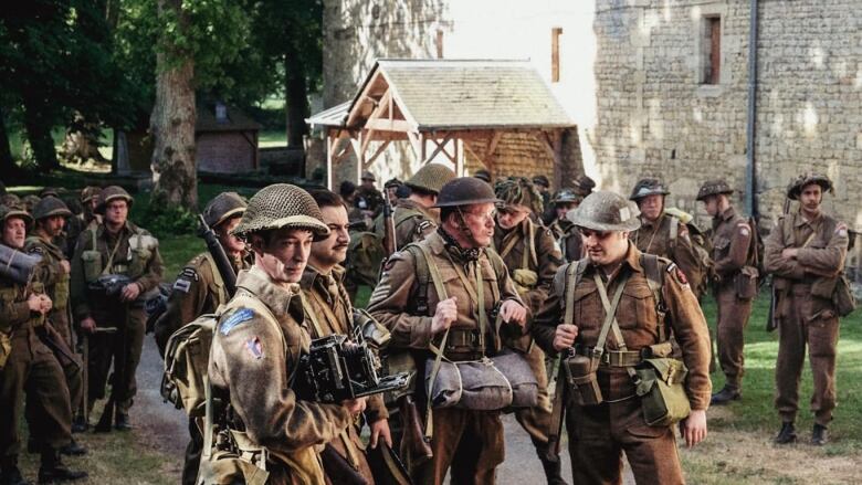 A group of men in second world war dress stand assembled near a stone building.