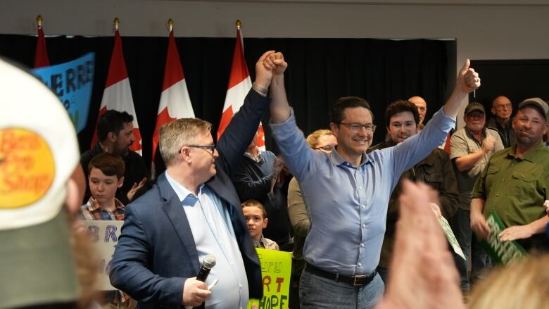 Two men hold their arms up in the air cheering as they address a crowd of supporters.