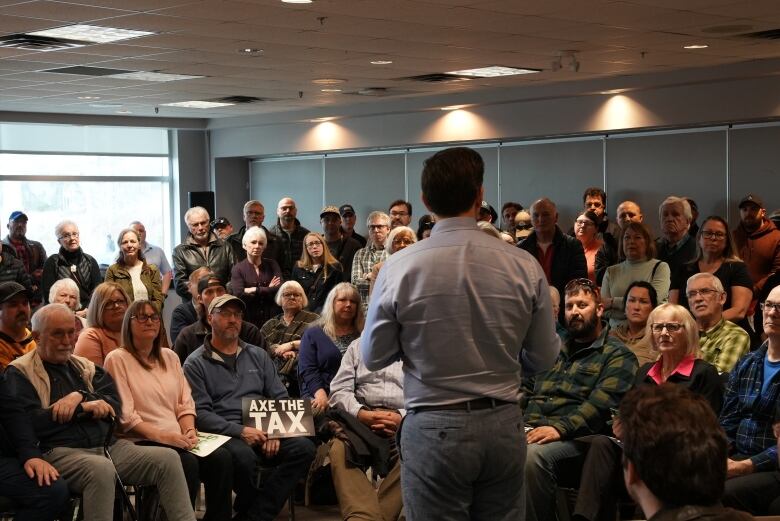 A crowd of people listen intently to a man wearing a dress shirt and grey pants.