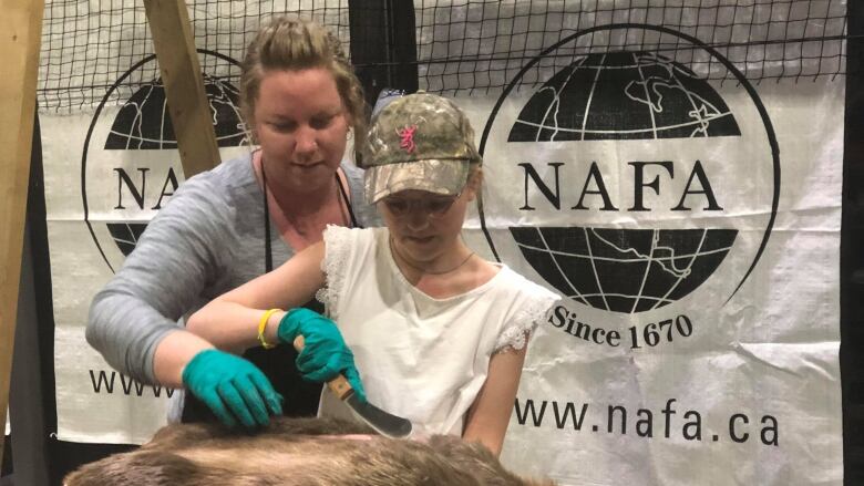 A woman and a youngster stand at a table, and the woman is showing the youngster how to remove the pelt from a beaver.