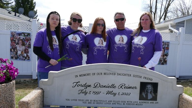 A family is pictured behind a bench engraved with a woman's name, photo and dates of her birth and death.