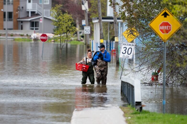 Two people in hip waders carry boxes out of a flooded part of a street.