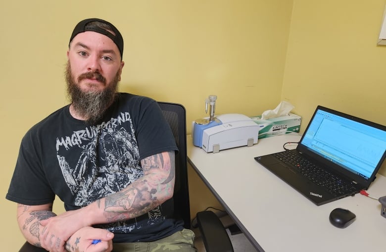 A man dressed in a Black T-shirt and backward ball cap sits facing the camera, next to him is a laptop and a white machine known as a Fourier-transform infrared (FTIR) spectrometer.