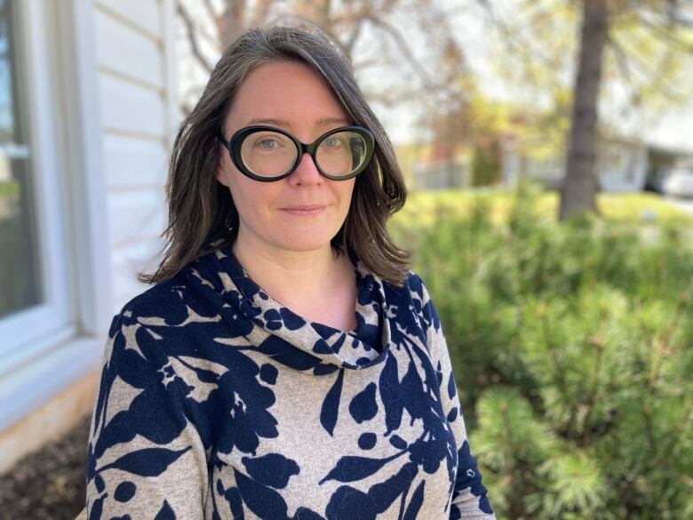 A woman with long dark hair and glasses stands outside in front of a house.