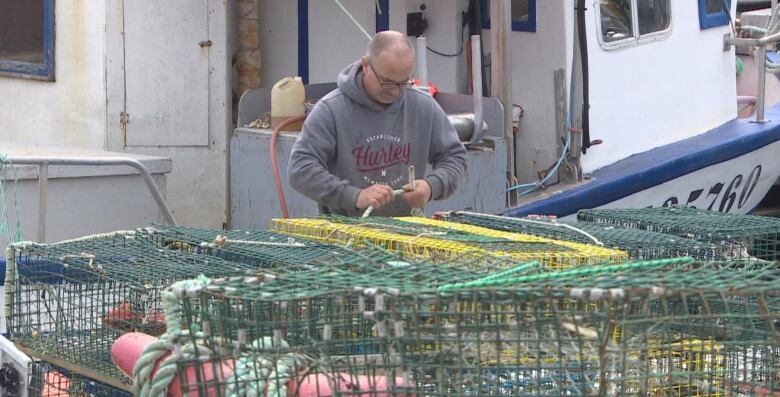 A man in a grey hoodie inspects lobster traps beside a fishing boat.