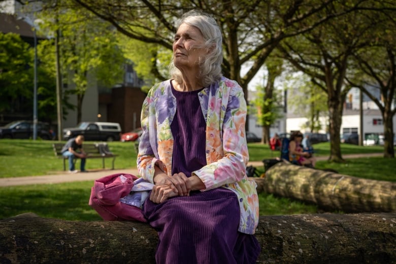 A woman with grey hair, wearing purple and a flower sweater, sits on a log