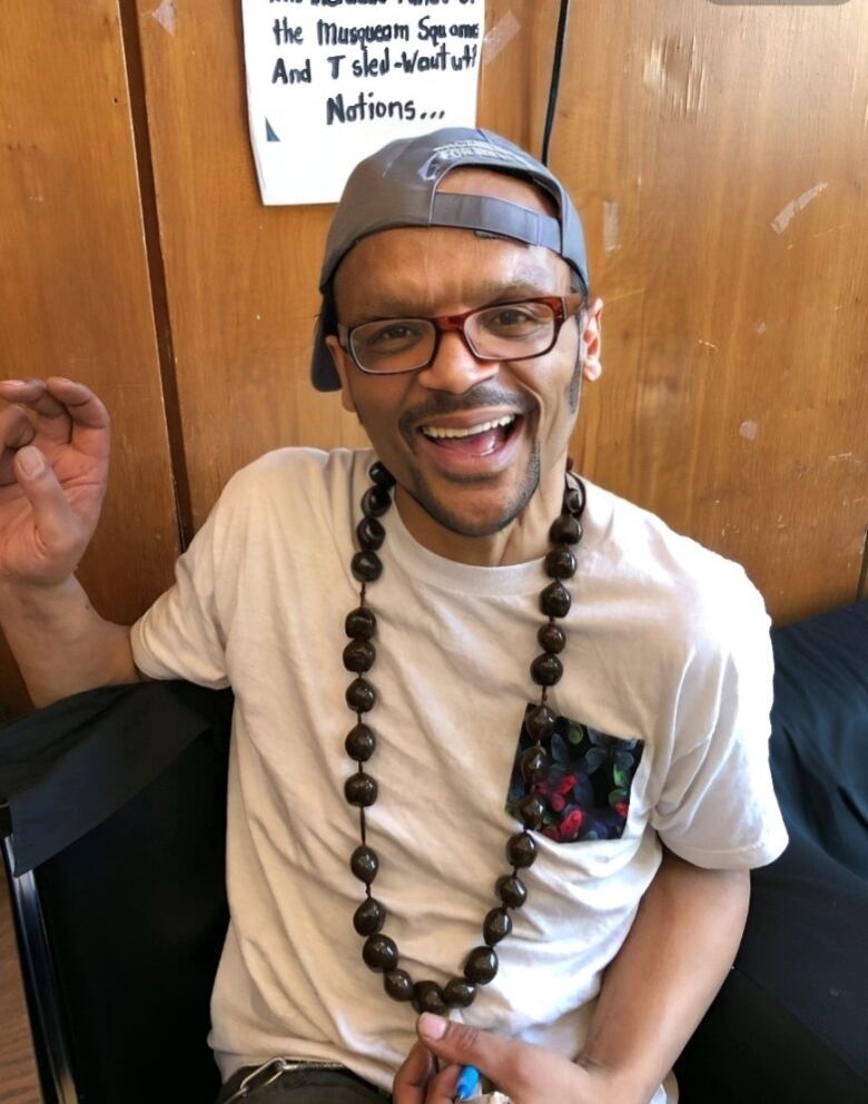 A man wearing a white T-shirt and a long beaded necklace smiles