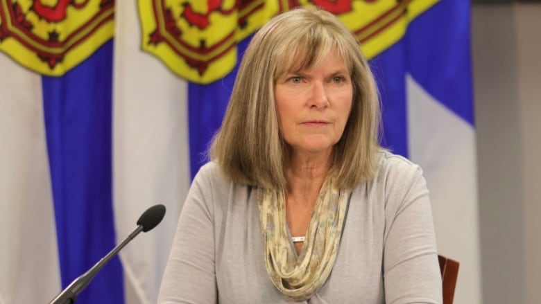 A woman sits in front of a microphone, with Nova Scotia flags visible in the background.