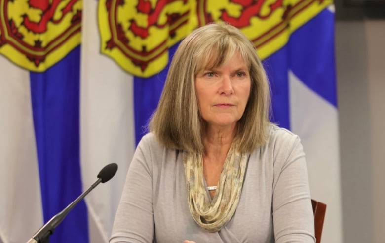 A woman sits in front of a microphone, with Nova Scotia flags visible in the background.