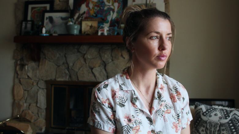 A woman sits in her living room, facing the camera, but looking out the window. In the background, art sits on a mantel above her fireplace.