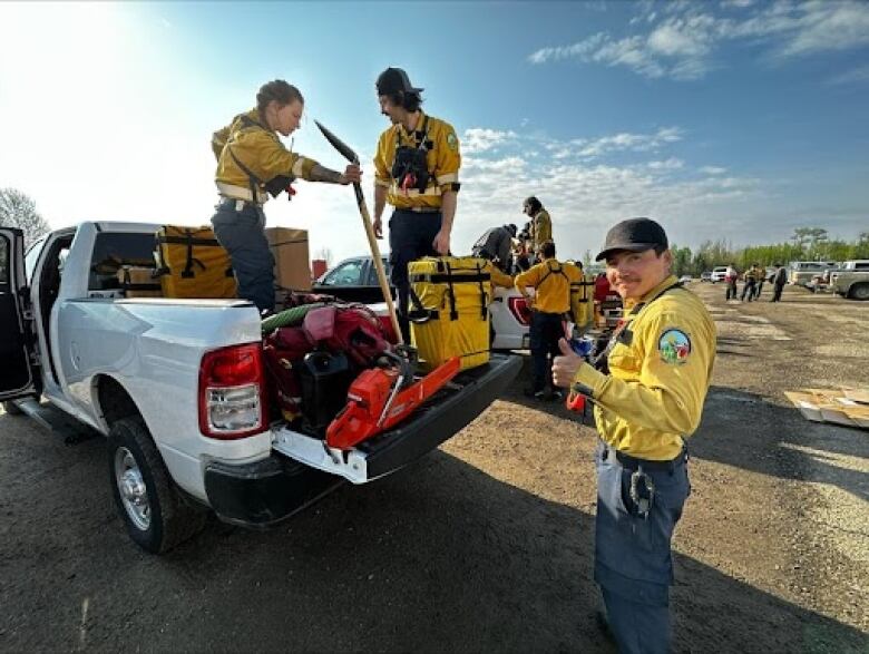 Two firefighters in yellow and blue clothing stand in the bed of a white pickup truck loaded with equipment. A third firefighter stands behind the truck.