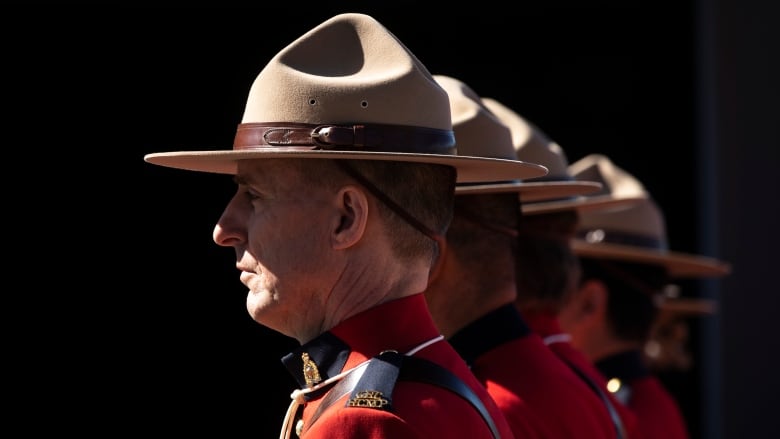 Profile picture from the shoulder up of a Mountie, facing left, wearing traditional red serge uniform. Other officers line up increasingly out of focus behind him.