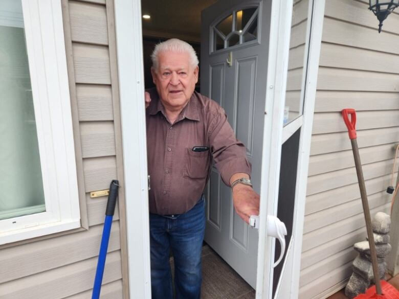 A white haired man with a brown shirt and jeans on stands holding the door to his house open