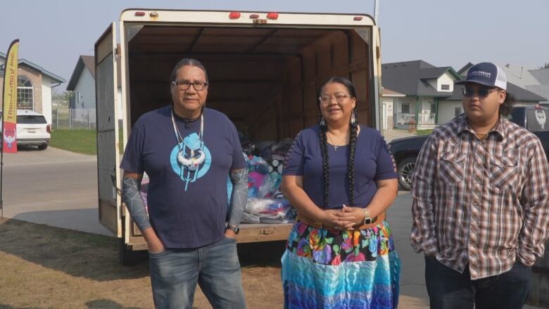 Three Indigenous people stand on a lawn, in front of a open trailer. The people are facing the camera. Inside the trailer is piles of bags filled with clothes, blankets and pillows. The trailer is parked in the driveway.