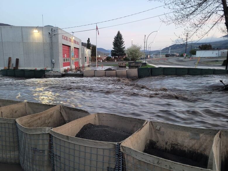 A river full of water with sandbags on the river bank and the Cache Creek fire hall in the background.