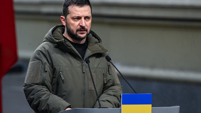 A man with a beard dressed in a khaki jacket stands at a microphone podium.