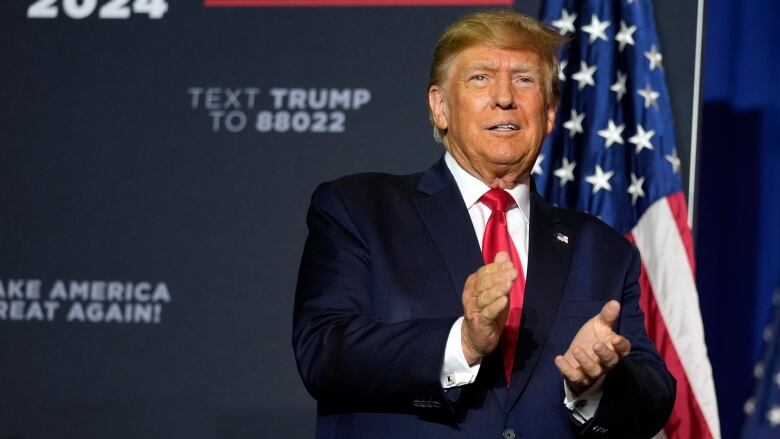 A person standing on a stage with a backdrop behind them claps their hands.