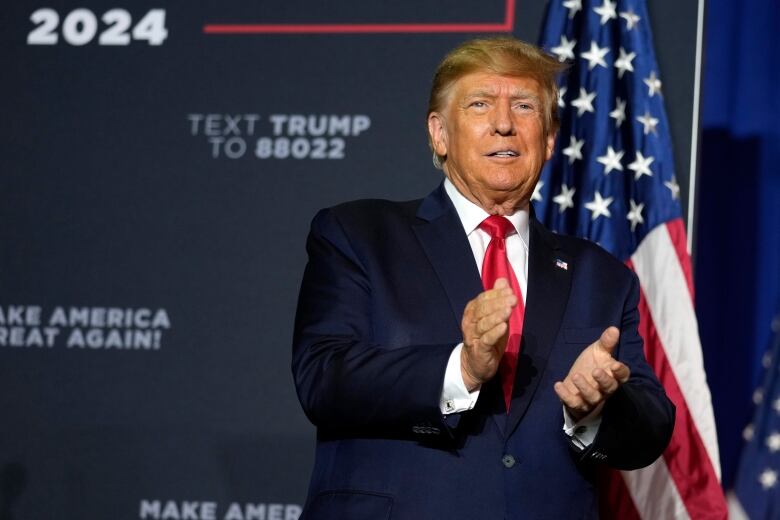 A person standing on a stage with a backdrop behind them claps their hands.