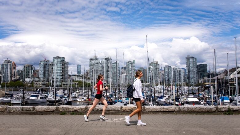 Two young women in profile, one walking, one jogging, on a seawall path in front of a marina with a city skyline of high-rises in the background.