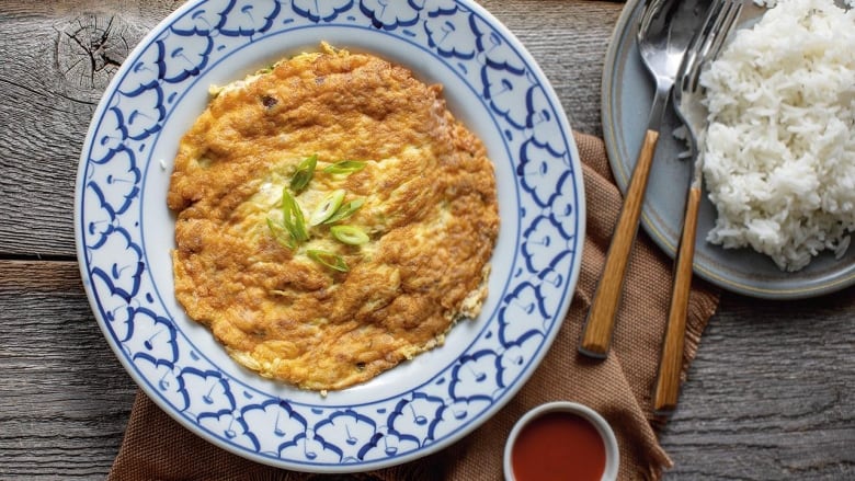 A bird's eye view of a plate setting with the main dish being golden brown omelet with sliced green onion on top for garnish. Beside it to the right is a slightly smaller dish with a spoon and fork and steamed rice. To the bottom is much smaller dish filled with a red dipping sauce.