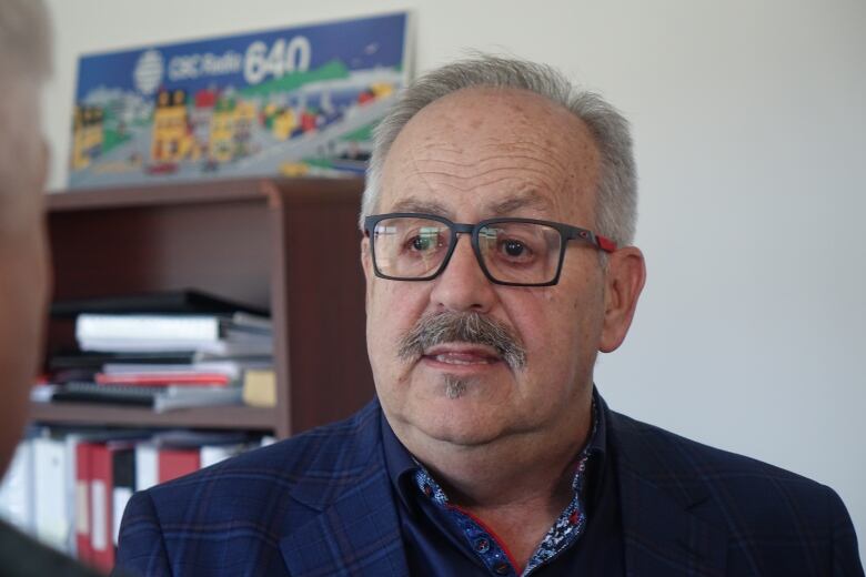 A man wearing a plaid blazer with grey and red glasses stands in an office.