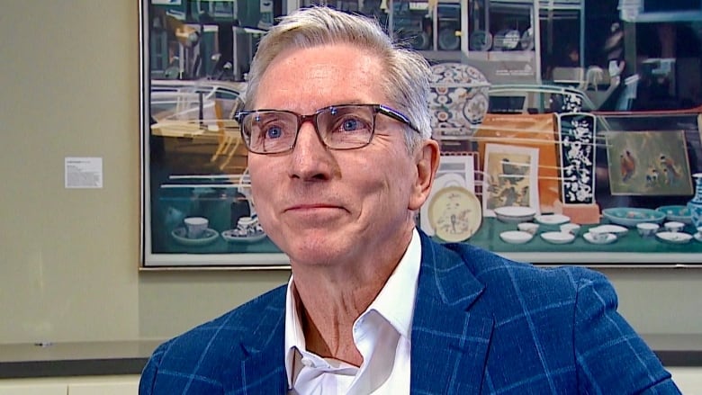 A man in a suit is sitting in a boardroom with a large painting hanging on the wall in the background.