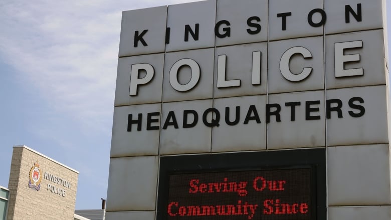 A big steel sign divided into more than 20 small squares sits in front of a grey brick building with the words Kingston Police and the service's logo next to it. The sky is blue with just wisps of cloud.