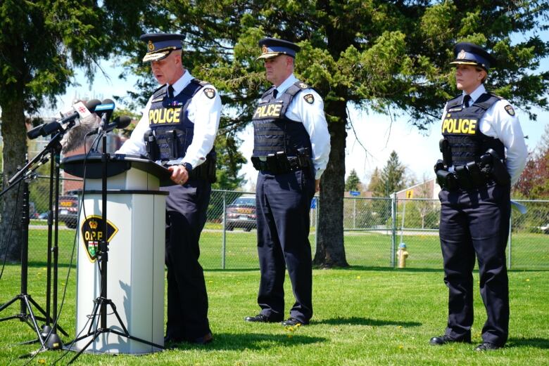 Three police officers stand on a lawn outside. The officer at the left speaks into microphones set up at a podium.