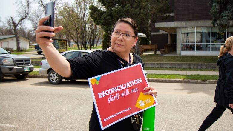 A woman takes a selfie while holding a sign saying 