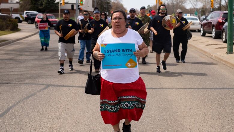 A woman wearing a ribbon skirt holds a sign saying 