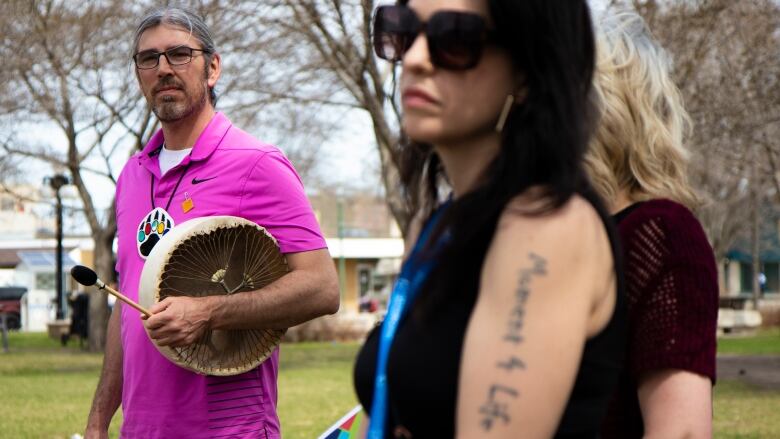 A man wears a pink shirt and holds an Indigenous drum.