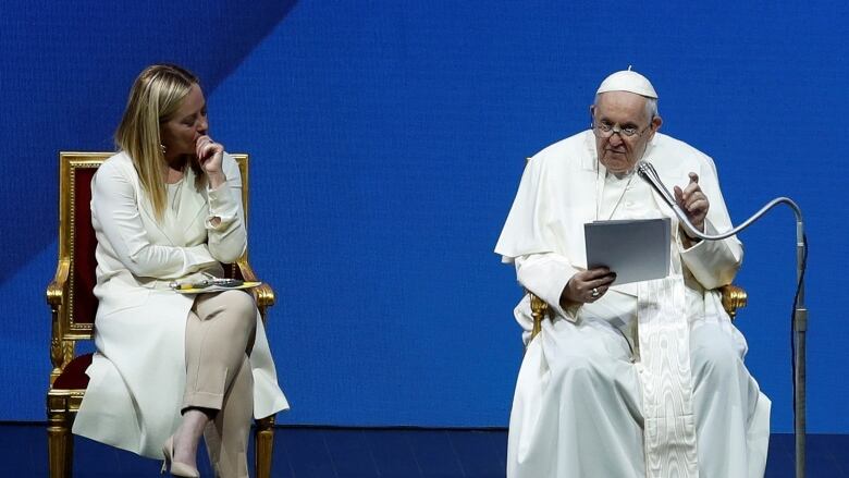 The pope is shown in religious garments speaking from papers he's holding while seated on a stage. To his right, a woman looks on.