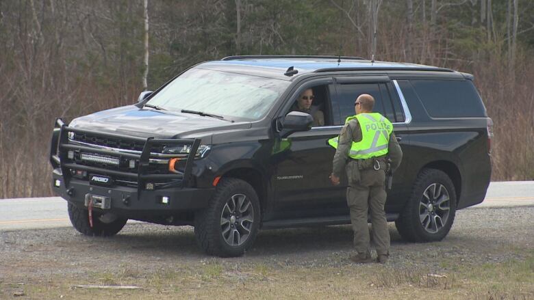 A man in uniform wearing a fluorescent police vest speaks with another officer who is inside a vehicle parked along a highway.