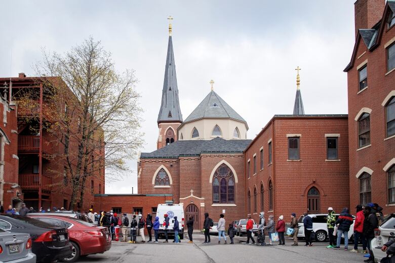 People form a long line in a parking lot. Red brick buildings are in the background and, farther back, church steeples. 