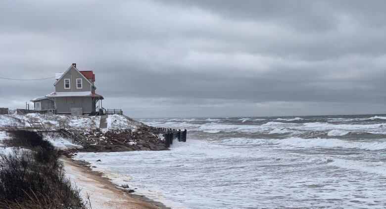 A house on a rocky hill by the beach with harsh waters in front of a grey, cloudy sky.