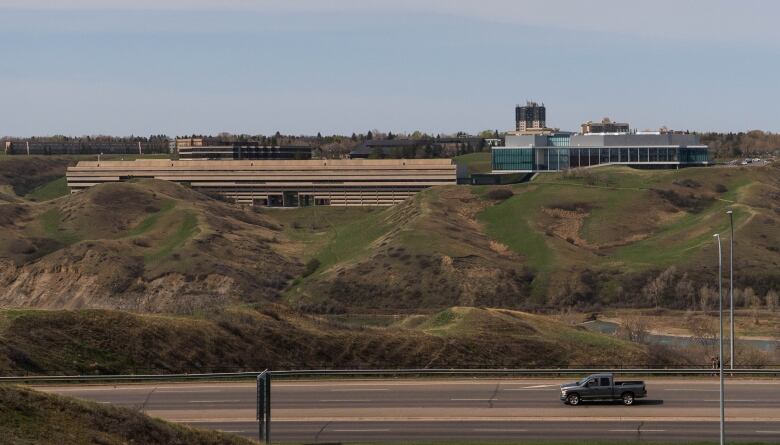 Two large concrete and glass buildings sit on top of green hills, with a truck driving past on a road in the foreground.