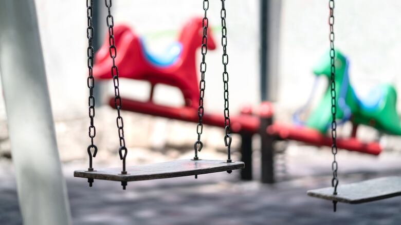 An empty swing is seen in the foreground with playground equipment in a blurred backgroun.