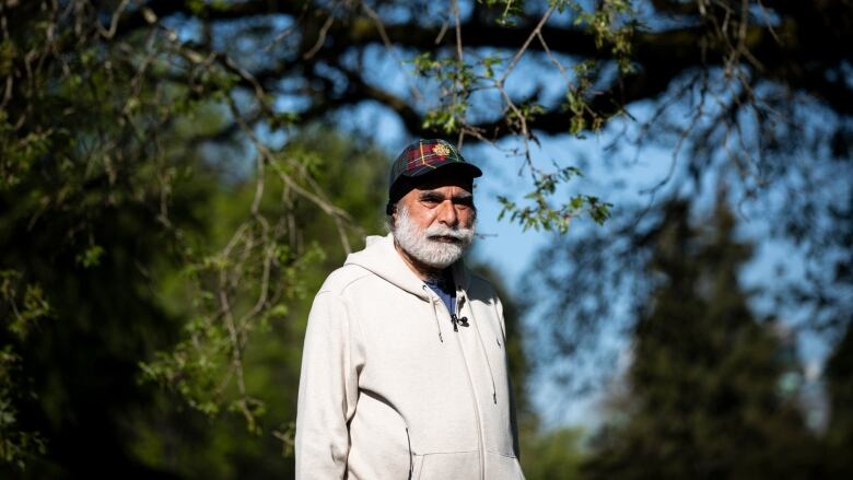 A bearded man wearing a white hoodie and a ballcap stands in the sun with trees behind him.