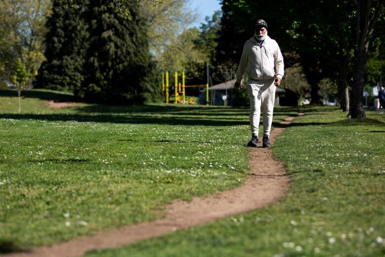 A man wearing a white sweatsuit and a ballcap walks along a path in a park.